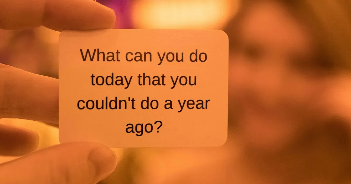 Hand holding a card that reads 'What can you do today that you couldn't do a year ago?' against a warm orange background
