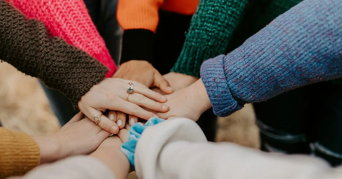 A group of hands stacked together in a circle, each wearing colorful knit sweaters, symbolizing team collaboration and partnership
