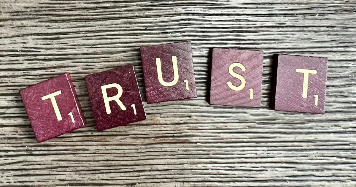 Dark red wooden letter tiles spelling "TRUST" arranged on a weathered gray wood surface