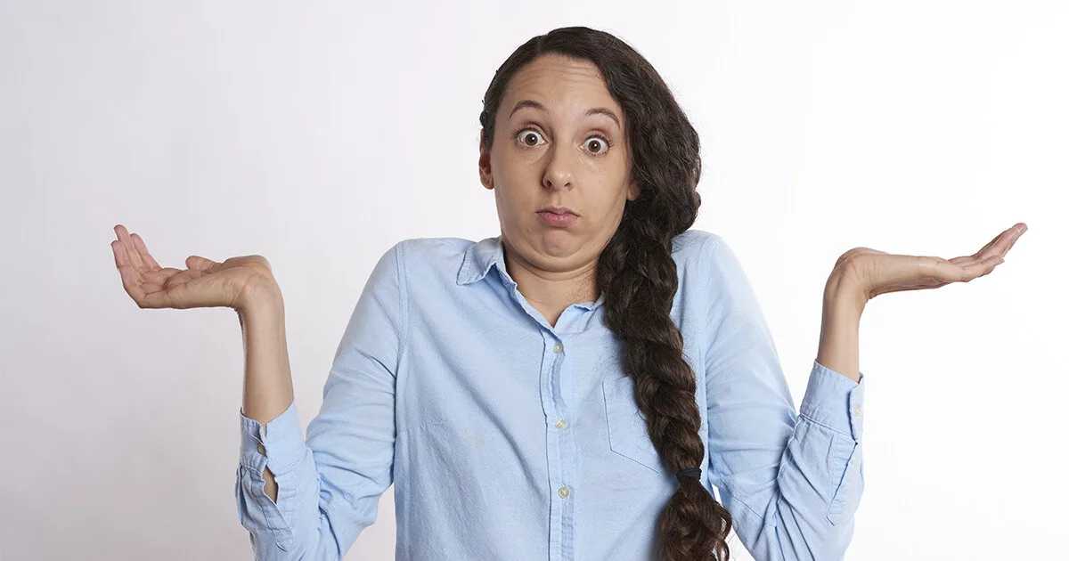 A woman in a blue button-down shirt shrugging with both palms up and a confused expression