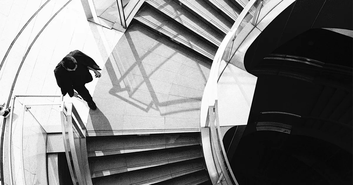 Black and white overhead photograph of a person descending a modern curved staircase with geometric shadows